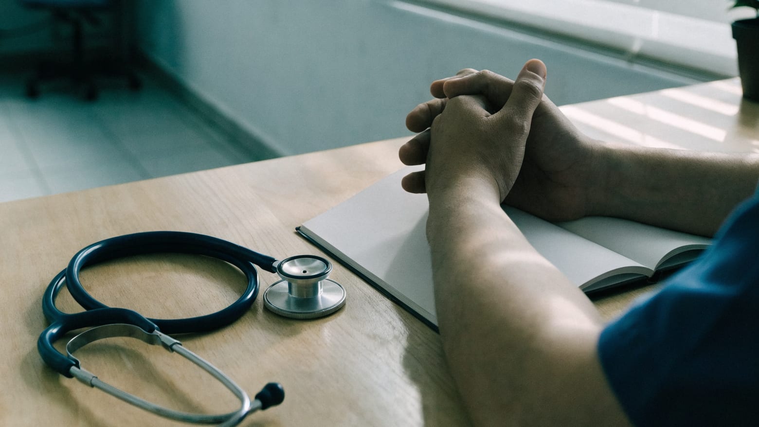 A doctor's clasped hands resting on an open notebook at the consultation-room desk, beside a stethoscope.